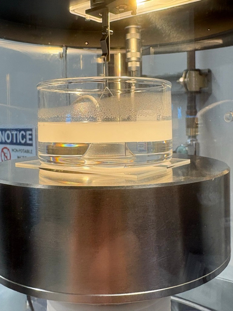 A glass vessel containing liquid on a circular metal platform beneath suspended fittings inside a laboratory instrument.