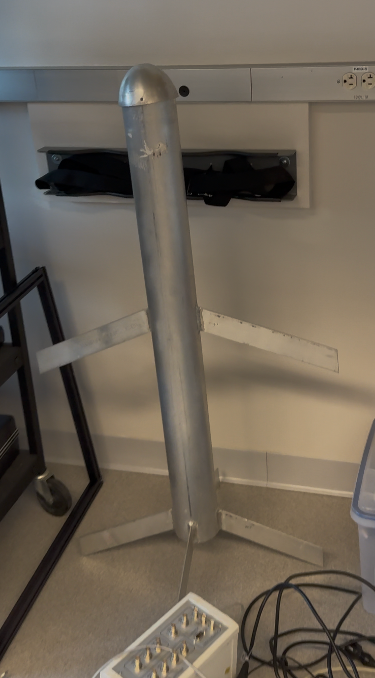 Photograph of a metallic cylindrical glider body with a rounded nose and fins, standing upright in the laboratory.