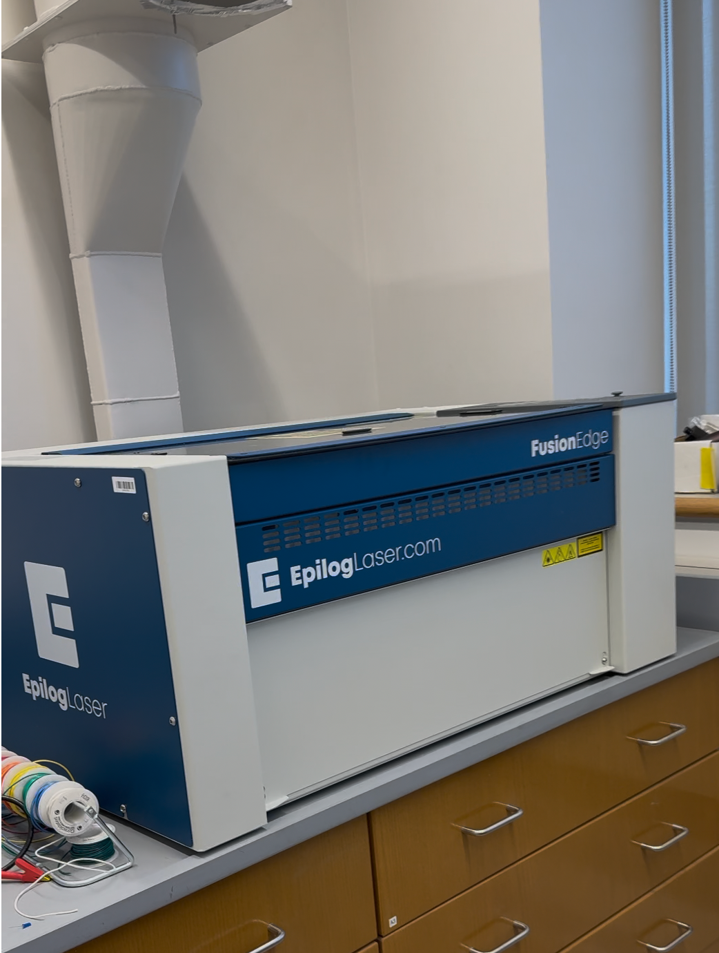 A blue-and-white enclosed laser cutter on a laboratory countertop, with a large ventilation duct behind it.