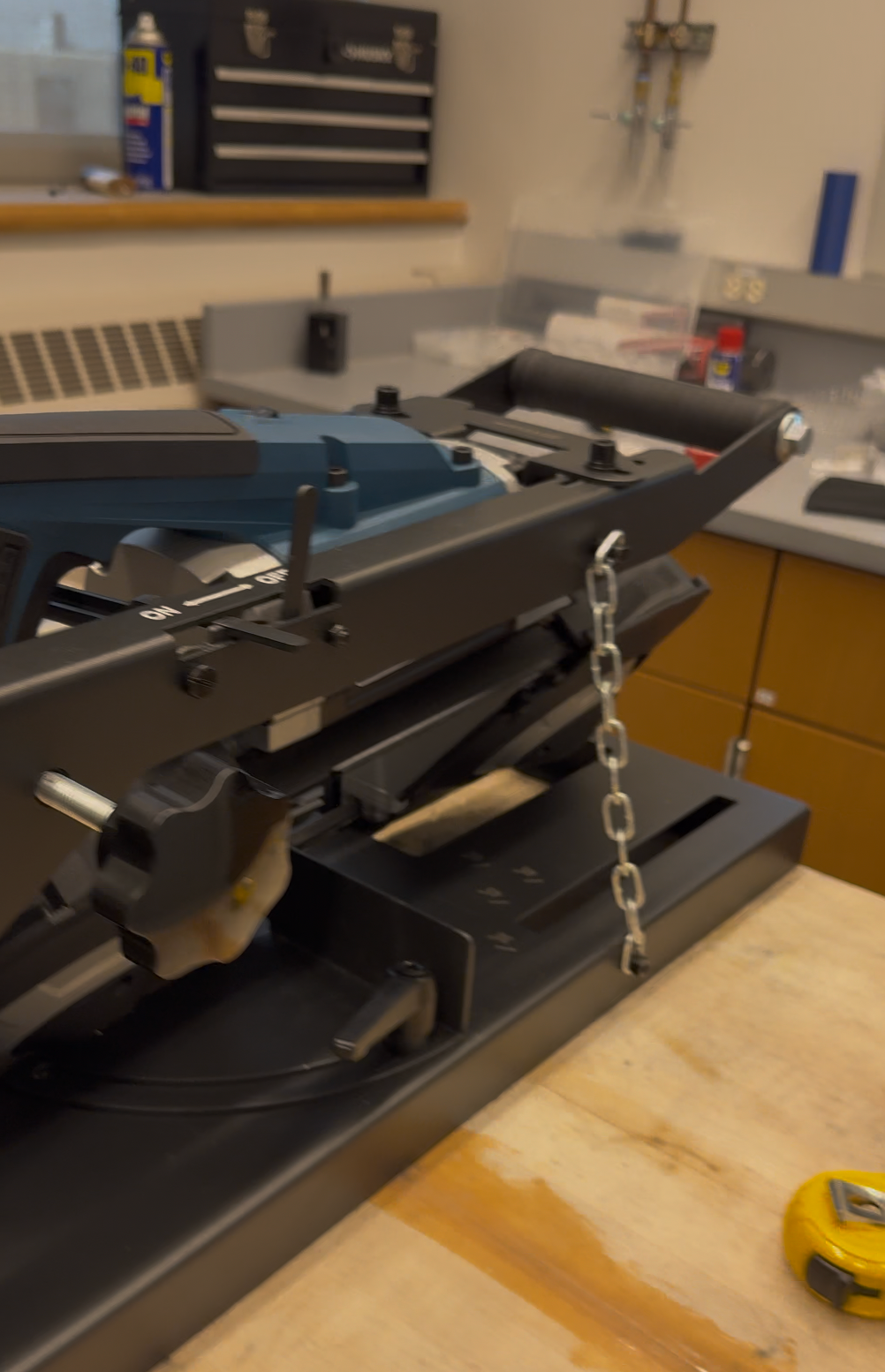 A benchtop saw with a blue-and-black upper assembly, adjustment knobs, a metal base, and a hanging chain.