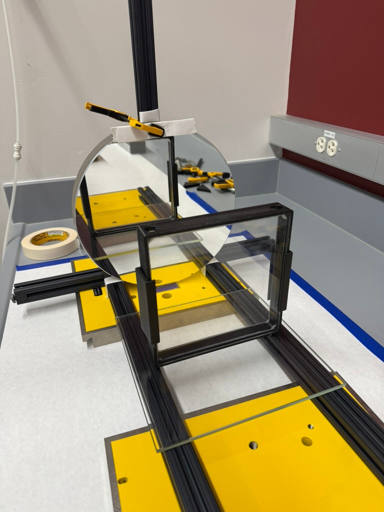 Schlieren setup with a circular mirror and a rectangular transparent panel mounted on black rails and yellow plates.